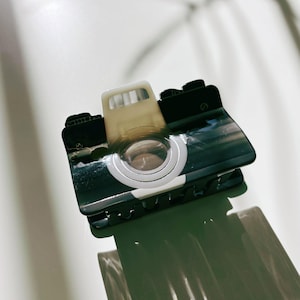 May include: Black and white camera-shaped hair clip with a white circle in the center. The clip has a textured surface and is made of plastic.