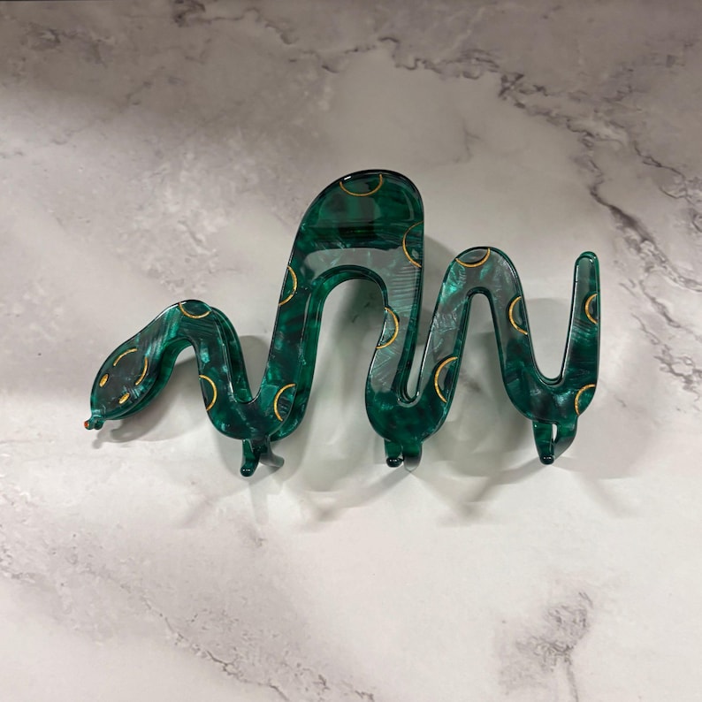 May include: A green and gold snake-shaped hair clip. The clip is made of a translucent material with a marbled effect. Gold crescent shapes are evenly spaced along the snake's body. The snake's head has small black eyes and a red dot.