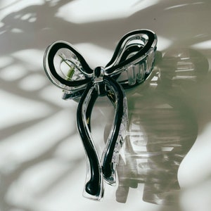 May include: A clear plastic hair claw clip in a bow shape with black accents. The clip has a spring mechanism and is casting a shadow on a white surface.