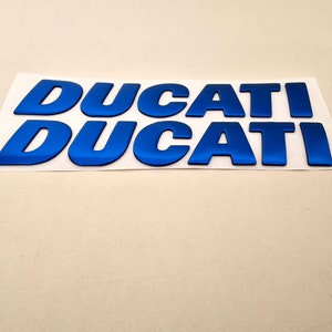 May include: Two sets of blue metallic "DUCATI" decals on a white backing. The letters are bold and slightly curved, with a reflective sheen. The decals are likely for a motorcycle or other vehicle.