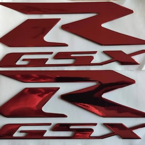 Puede incluir: Dos juegos de pegatinas de vinilo rojo brillante con las letras "GSX" y una "R" estilizada en una pegatina de vinilo rojo brillante. Las pegatinas están sobre un fondo blanco.