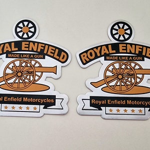 May include: Two silver Royal Enfield motorcycle stickers with a black and gold cannon design. The text on the stickers reads "Royal Enfield" and "Made Like A Gun". The stickers also include the text "Royal Enfield Motorcycles" and five stars.