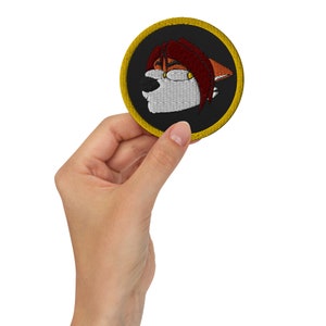 May include: A round embroidered patch with a black background and a yellow border. The patch features a cartoon fox with red hair, white fur, and black eyes.