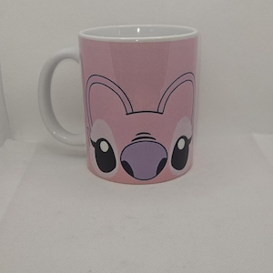 May include: A pink ceramic mug with a cartoon image of Stitch's face. The mug has a white handle and a white rim.
