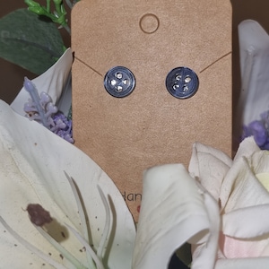May include: A pair of black stud earrings with a circular design featuring four small silver studs. The earrings are displayed on a brown card with the word "Handmade" visible.