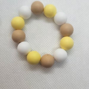 Puede incluir: Un brazalete hecho de cuentas de silicona en colores amarillo, blanco y marrón. Las cuentas son redondas y tienen un acabado mate.