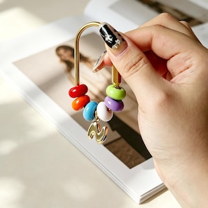 Pode incluir: Um suporte para berloques de metal dourado em forma de U com contas lisas e coloridas em vermelho, laranja, azul, branco, roxo e verde. Um berloque dourado em forma de letra N pende da parte inferior da estrutura de metal em forma de U.