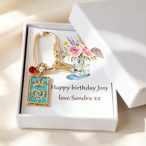 May include: A gold-toned keychain featuring a rectangular turquoise charm with a crown and heart design, a red crystal, an evil eye bead, and a letter N charm. It is presented in a white box with a floral illustration and the text Happy birthday Josy love Sandra xx.