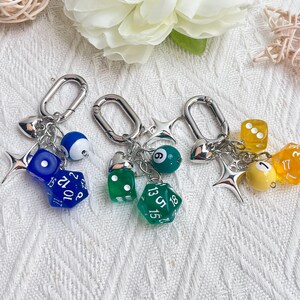 May include: Three keychains with silver clasps, each featuring a different combination of charms. One has a blue die, a blue and white billiard ball, and a star. Another has a green die, a green billiard ball, and a star. The third has a yellow die, a yellow billiard ball, and a star.