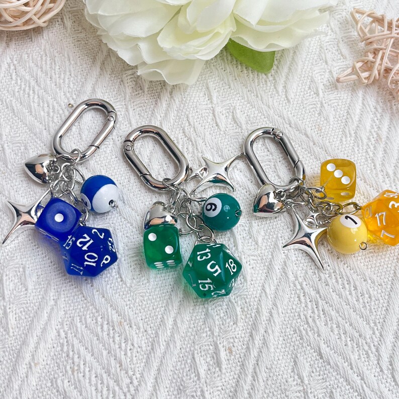 May include: Three keychains with silver clasps, each featuring a different combination of charms. One has a blue die, a blue and white billiard ball, and a star. Another has a green die, a green billiard ball, and a star. The third has a yellow die, a yellow billiard ball, and a star.