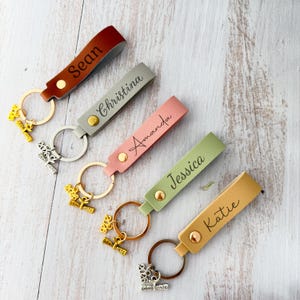 May include: A collection of personalized keychains in various colors: brown, gray, pink, green, and yellow. Each keychain features a name and a graduation-themed charm. The keychains are displayed on a light-colored wooden surface.