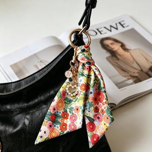 Puede incluir: Un charm de bufanda con estampado floral con un anillo dorado y un charm en forma de corazón con la letra "N", colgando de un bolso negro. La bufanda presenta un diseño floral colorido con flores rojas, rosas, amarillas y azules.
