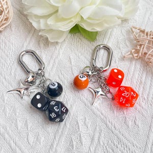 May include: Two keychains with silver-tone clasps. One keychain features a black eight-ball, black dice, and a star charm. The other has an orange ball, red dice, and a star charm. The background is a white, textured surface.