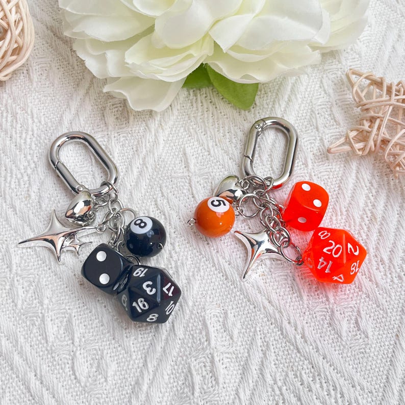 May include: Two keychains with silver-tone clasps. One keychain features a black eight-ball, black dice, and a star charm. The other has an orange ball, red dice, and a star charm. The background is a white, textured surface.