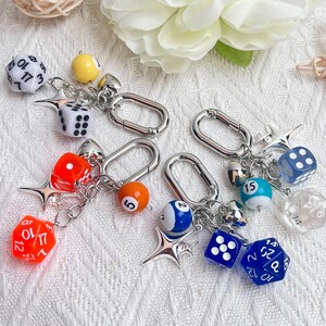 May include: Keychains with dice and billiard ball charms. Each keychain has a silver clasp and features a variety of dice, including D20s and D6s, and small billiard balls. The dice are in various colors, including white, orange, and blue.