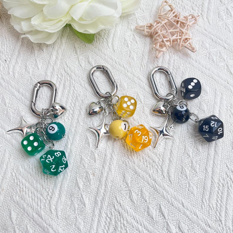 May include: Three keychains with silver clasps, each adorned with dice, star charms, and heart pendants. One keychain features green dice and a pool ball. Another has yellow dice and a yellow ball. The third has black dice and a pool ball.