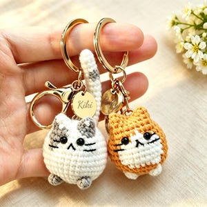 May include: Two round crocheted cat keychains with gold-toned metal rings and clasps. One cat is white with gray patches, and the other is orange and white. Each features black bead eyes, embroidered whiskers, and a small round gold tag.