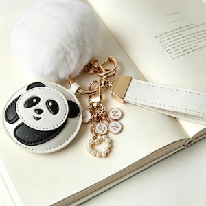 May include: A keychain featuring a white faux fur pom-pom, a circular white and black panda face charm, and a white leather strap. The gold-toned hardware includes a heart-shaped charm with small white beads and circular charms with the letters L, E, N, N.