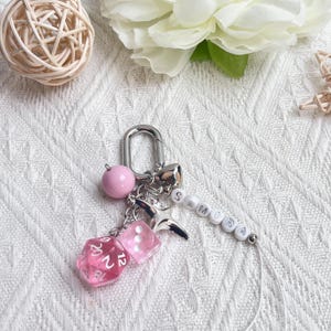 May include: A silver keychain with a pink bead, a star charm, and two pink dice. The keychain also features the name "SAMIRA" spelled out in white beads. A small silver bell is also attached.
