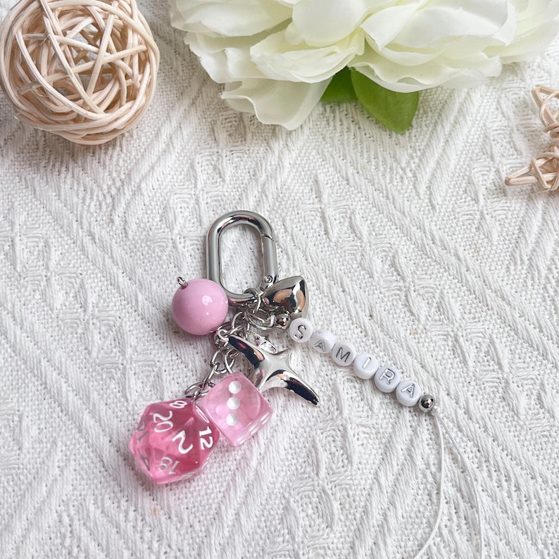 May include: A silver keychain with a pink bead, a star charm, and two pink dice. The keychain also features the name "SAMIRA" spelled out in white beads. A small silver bell is also attached.