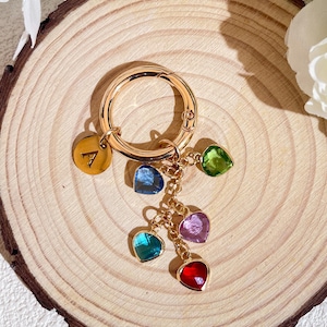 Puede incluir: Llavero dorado con un dije con la letra A y cinco dijes colgantes en forma de corazón con piedras preciosas de diferentes colores: azul, verde, rosa y rojo.