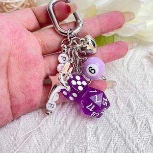May include: A silver keychain with multiple charms, including a purple eight ball with the number 6, two purple dice, a silver heart, a star, and letter beads spelling "PR". The keychain is attached to a silver carabiner.