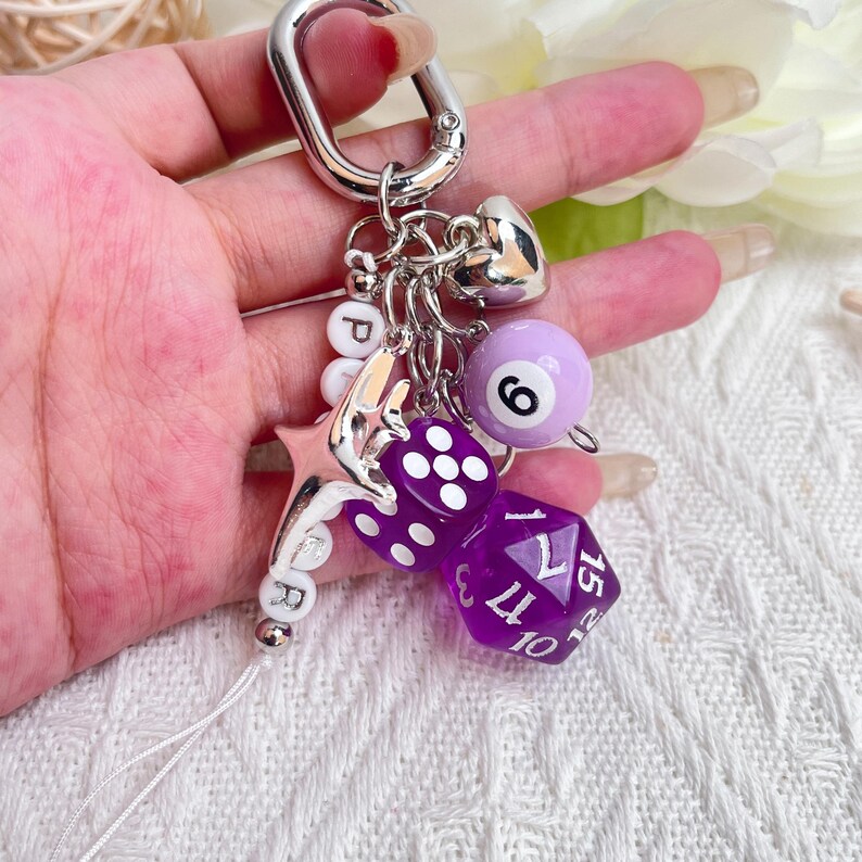 May include: A silver keychain with multiple charms, including a purple eight ball with the number 6, two purple dice, a silver heart, a star, and letter beads spelling "PR". The keychain is attached to a silver carabiner.