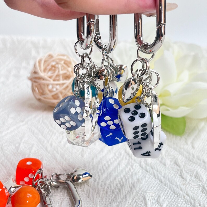 May include: Silver-toned keychain with multiple dice charms in various colors, including blue, white, and orange. The dice have white dots. The keychain is suspended against a soft, white background.