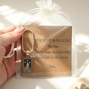May include: A gold-toned oval carabiner keychain featuring a rectangular black tarot card charm with a gold figure, a small red bead, and a letter N charm. It is packaged in a sheer white organza bag with a card reading FOR HOPE & HEALING.