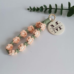 Pode incluir: Um porta-chaves de macramé feito à mão com detalhes de flores cor de pêssego e um cordão verde. O porta-chaves inclui uma etiqueta redonda com o nome "Monica" e a inicial "M". O hardware é dourado.
