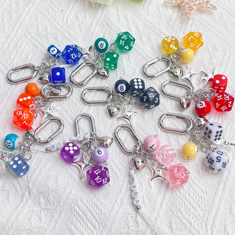 May include: A collection of colorful dice keychains. Each keychain features a silver clasp, a variety of dice in different colors, and decorative charms. The dice include D6, D12, and D20, with numbers visible. Some keychains have additional beads and star charms.