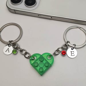 Può includere: Due portachiavi argentati collegati da una catena, con un ciondolo a forma di cuore verde fatto di mattoncini. Ogni portachiavi ha un ciondolo rotondo con una lettera e una gemma colorata. Uno ha la lettera "A" e una gemma verde, l'altro ha la lettera "E" e una gemma rossa.