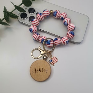 May include: A red, white, and blue beaded phone charm with a gold key ring and a wooden charm with the name "Ashley" engraved on it.