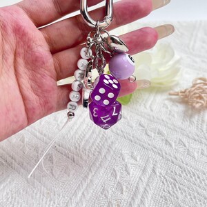 May include: A silver keychain with multiple charms. Charms include two purple dice, a purple billiard ball with the number 6, and a string of white letter beads spelling out a name. The keychain is attached to a silver clasp.