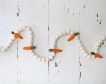 May include: A white felt ball garland with orange felt carrots attached. The garland is strung in a wavy pattern on a white wooden background.