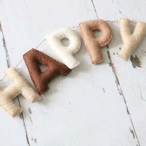 May include: A felt banner with the word "HAPPY" spelled out in brown, beige, and white letters. The letters are strung together with twine.