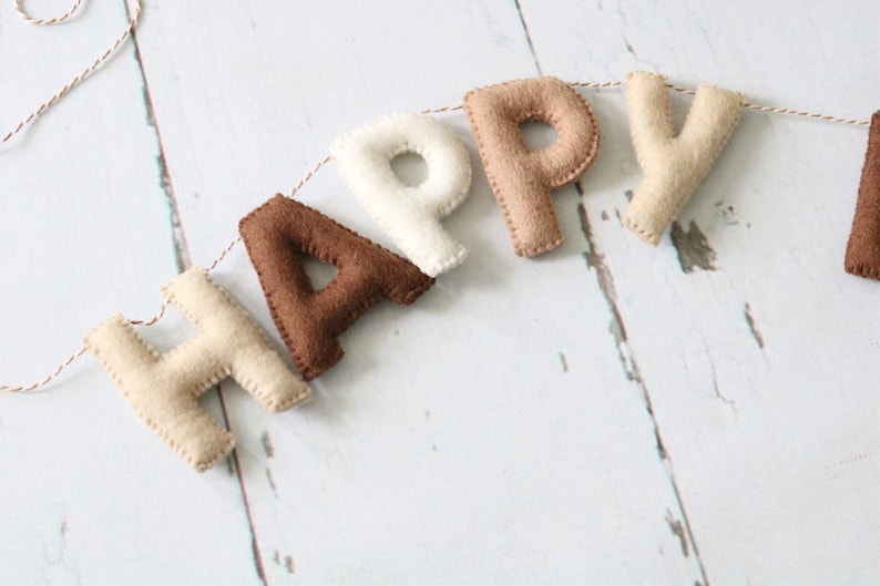 May include: A felt banner with the word "HAPPY" spelled out in brown, beige, and white letters. The letters are strung together with twine.