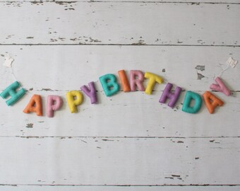 May include: A colorful felt banner spelling "HAPPY BIRTHDAY" in various colors, including teal, orange, pink, yellow, and purple. The letters are strung together with a white string, set against a white wooden background.