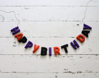 May include: A felt letter garland spelling out the words HAPPY BIRTHDAY in a repeating sequence of purple, orange, and black colors. The individual stuffed letters are strung together on a thin dark string, creating a festive hanging banner.
