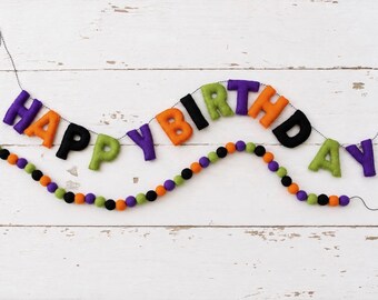 May include: A felt banner spelling out HAPPY BIRTHDAY in purple, orange, black, and lime green letters strung on a thin black cord, accompanied by a separate garland of alternating felt balls in the same color palette of purple, orange, black, and lime.