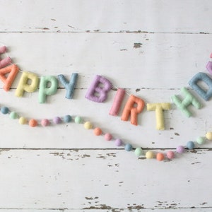 May include: A pastel-colored felt banner spelling "HAPPY BIRTHDAY" with matching felt ball garland. The letters are in shades of pink, orange, yellow, green, and blue, stitched onto a white string. The garland features pastel-colored felt balls.