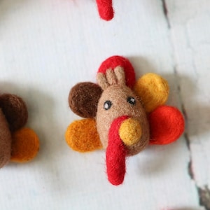 May include: A set of five felt turkey ornaments in various shades of brown, orange, red, and yellow. Each turkey has a brown felt body, a red felt wattle, and a yellow felt beak.