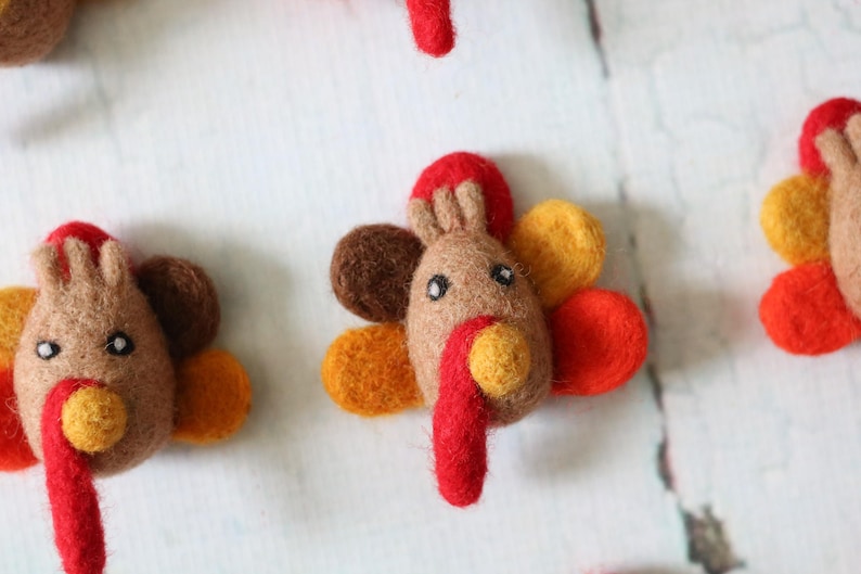 May include: A set of five felt turkey ornaments in various shades of brown, orange, red, and yellow. Each turkey has a brown felt body, a red felt wattle, and a yellow felt beak.