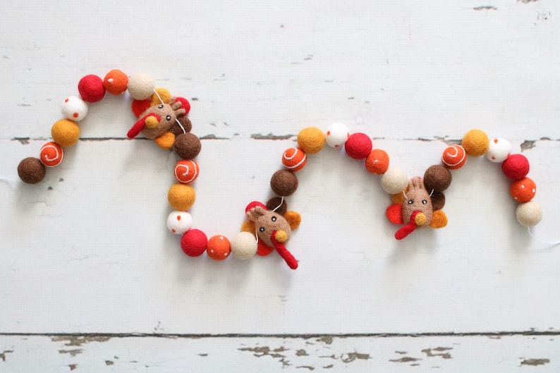 May include: A garland of felt balls in orange, red, white, and brown with three felt turkey decorations. The garland is strung on a white string and is perfect for Thanksgiving decor.