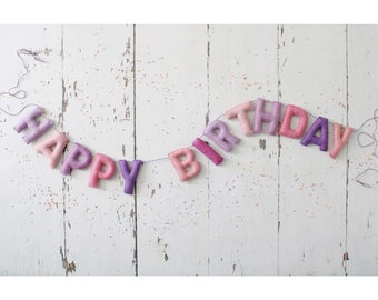 May include: A felt banner with the words "Happy Birthday" in purple, pink, and white. The banner is strung on a white string and is hanging against a white wooden background.