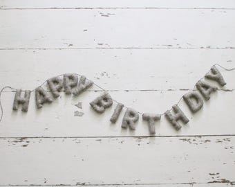 May include: A gray felt banner spelling "HAPPY BIRTHDAY" strung on a thin black string. The letters are puffy and textured, set against a white, weathered wooden background. A festive decoration for a birthday celebration.
