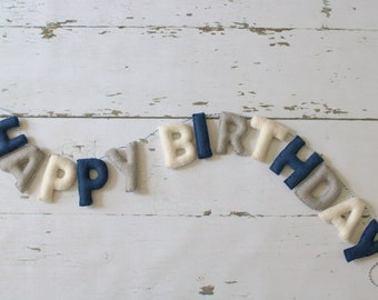 May include: A felt banner spelling "HAPPY BIRTHDAY" in navy blue, gray, and off-white letters. The letters are strung together with a thin, gray and white string, and are set against a white, distressed wooden background.