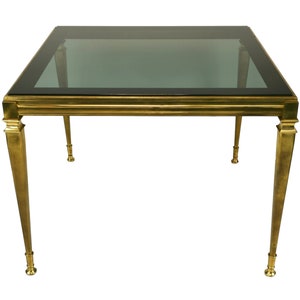 May include: A square coffee table with a dark glass top and a brass-colored metal frame. The legs are tapered and fluted, with decorative detailing at the top and bottom. The table is a classic design.