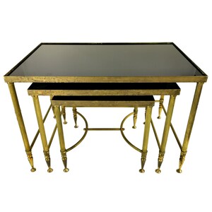 May include: A set of three nested tables featuring rectangular black glass tops and distressed gold-toned metal frames. Each table has four fluted legs with tapered feet and curved metal supports connecting the legs for structural stability.
