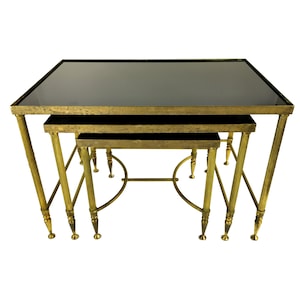 May include: A set of three nesting tables. Each table has a black glass top and a brass-coloured metal frame. The largest table has a rectangular top. The legs feature a fluted design.